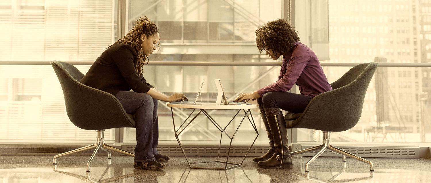 Two women sitting at a small circular table while typing on their laptops