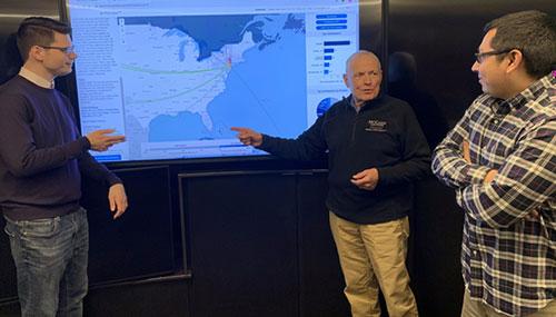Three men looking at a map on a large TV monitor.